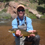 Man on one knee holding spotted brown trout with South Platte River in background
