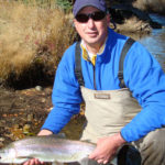 Man on knee holds large and healthy rainbow trout alongside idyllic river