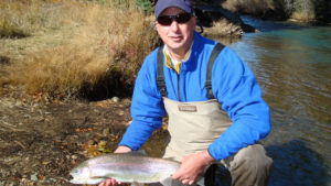 Man on knee holds large and healthy rainbow trout alongside idyllic river