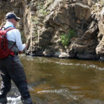 Man wearing red backpack wading in stream with bent fly rod and a rocky backdrop.