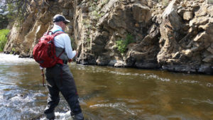 Man wearing red backpack wading in stream with bent fly rod and a rocky backdrop.