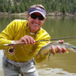 Fly fishing angler holds a brown trout with one hand and fly rod with the other