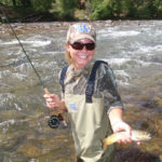 College girl in waders poses with brown trout and fly rod standing in swift river