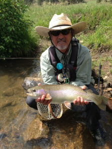 A rainbow trout is held streamside by a Denver fly fishing angler with a goatee and western hat