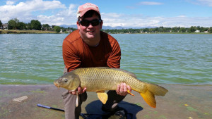 Smiling angler holding carp caught fly fishing in a lake, with mountains in the background.