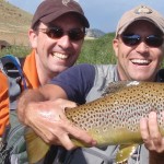 Two smiling friends kneeling along the Dream Stream in Colorado, one is holding a large brown trout.