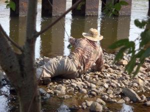 Fly fishing guide Chris Galvin lays on his side on the rocky edge of a river extending his fly rod to present to wary carp