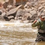 Man in fly fishing vest and waders prepares to net a Colorado River trout